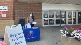 A woman in a striped shirt holds up a clipboard with voter information on it behind a table that says "Iowa Asian Alliance." A sign to her right says "Register to Vote Here." Grocery store sliding doors are behind her and flowers are to her left.