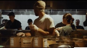 Several people stand in a food prep room, wearing hats and gloves. They are assembling free meals for a Thanksgiving event.