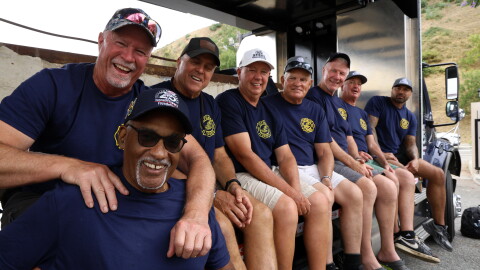 A row of men in blue T-shirts sit on a truck bed