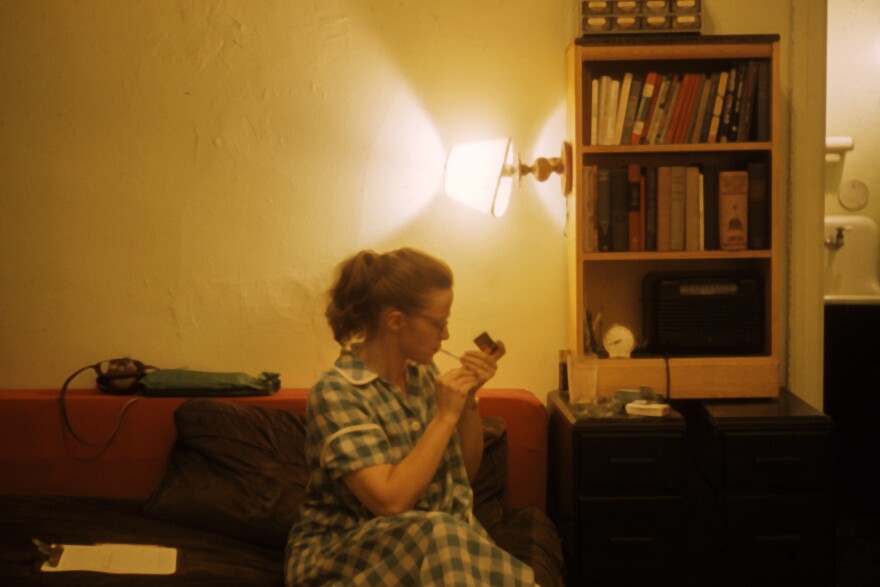 Connie Converse sits on a couch in her apartment while lighting a cigarette. 