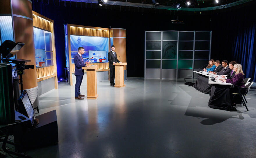 Two men in suits stand at podiums in front of a panel of five people at a TV studio