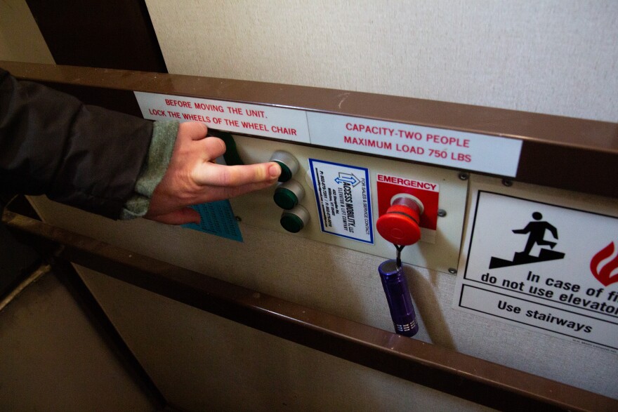 A photo of a light-skinned hand pushing a button. Around the hand are signs reading "Before moving the unit, lock the wheels of the wheel chair," "capacity two people maximum load 750lbs" and "in case of fire do not use elevator use stairways." There's also a red emergency button.
