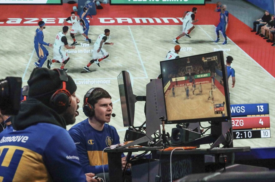 A giant monitor shows play as Warriors Gaming Squad teammates Charles "CB13" Bostwick, center, from New York, and his teammate Alexander Reese, left, from Milwaukie, Or., react to scoring during the NBA 2k League (NBA2KL) professional esports playoffs, Wednesday, July 24, 2019, in Queens borough of New York. The teams kicked off day one of the NBA2KL playoffs with Blazers on the losing 67-45. (AP Photo/Bebeto Matthews)