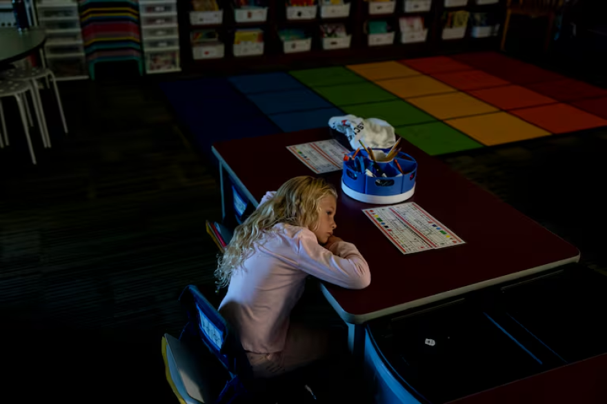 A kindergartener at a table in a darkened room with her head on the table.