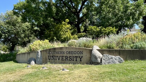 Southern Oregon University is etched into a stone wall. Behind it are trees and flowers. 