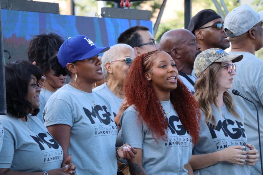 This photograph captures members of the Making Gay Music Famous organization standing together during an outdoor event, wearing matching gray shirts with the group's logo. The group is known for advocating LGBTQ+ rights and promoting visibility through music and public events.