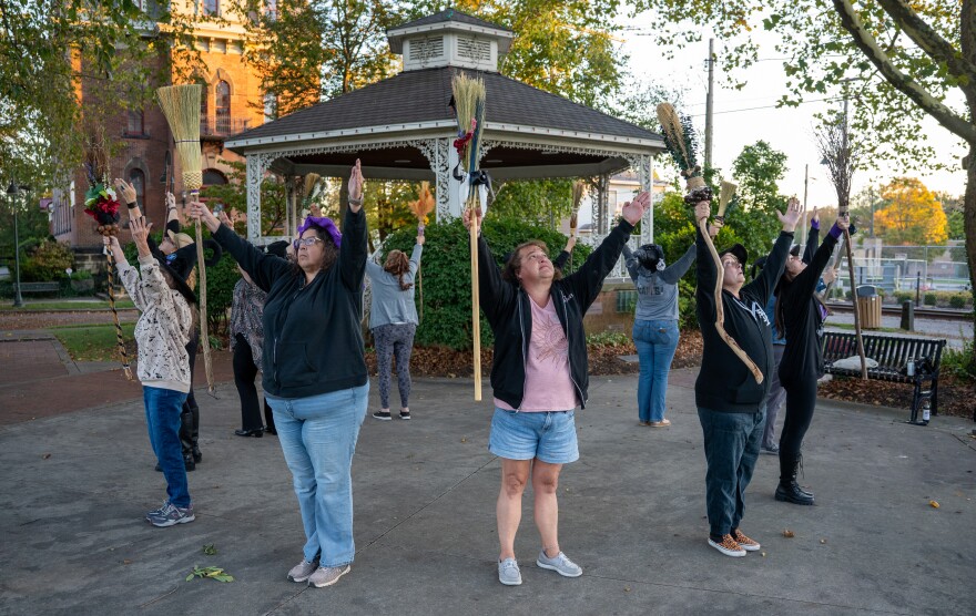 Group of women in a circle dressed as witches hold brooms skyward, in front of a gazebo in a public park.