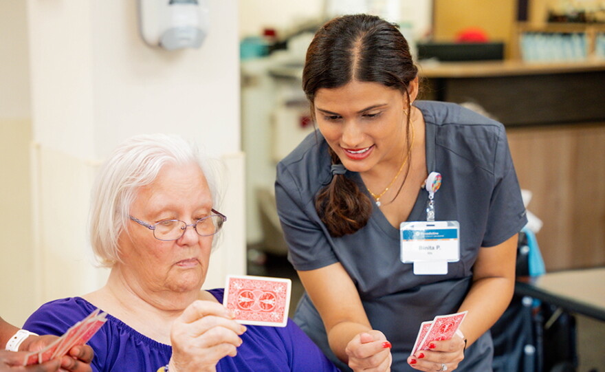A nurse in scrubs leans over while holding playing cards next to a nursing home resident, also holding playing cards.