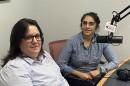 Two women in business casual clothing sitting in a recording studio with a microphone between the two of them.