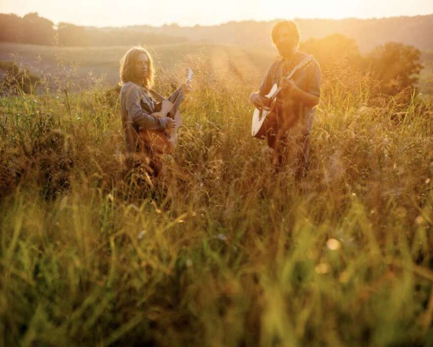 Carl Broemel and Tyler Ramsey are holding guitars while standing in a field of tall grass with the sun setting.