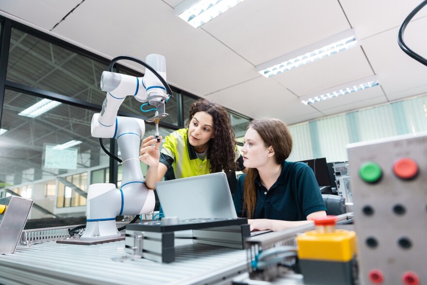 Engaged young woman student engineer with another woman learning about robot technology with a robotic arm