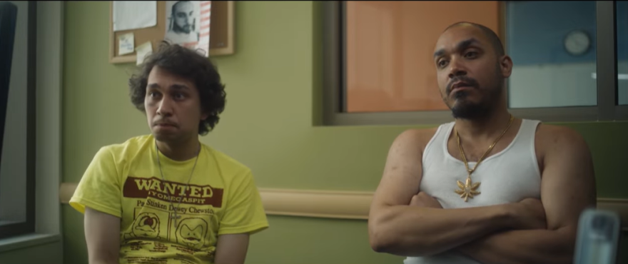 Alex sits in a yellow tee shirt with a red design and Dylan sits in a white cut off shirt and flashy gold chain.
