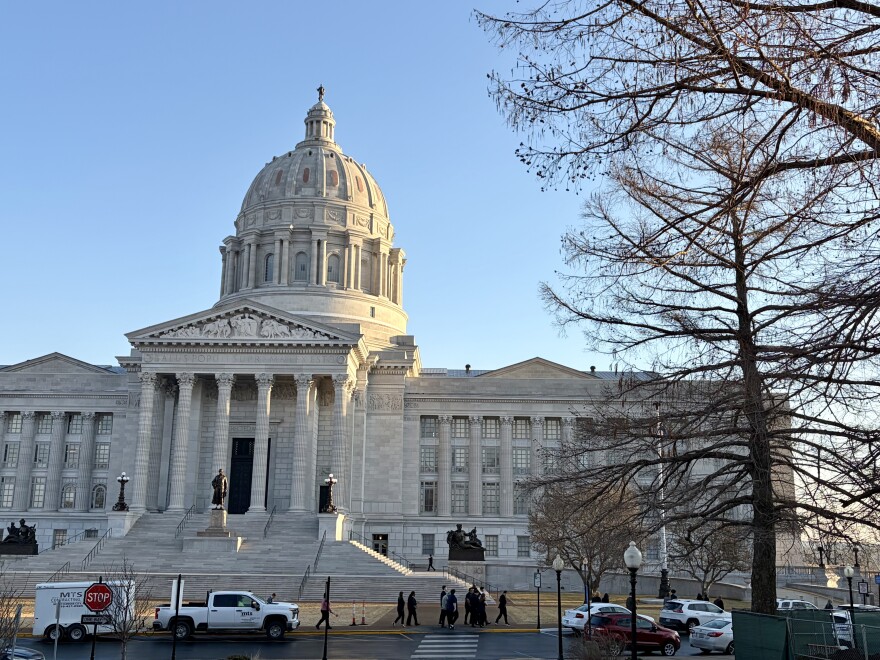 A photo of the exterior of the Missouri State capitol building. Vehicles are parked in front of the domed structure, people walk on the sidewalk. 