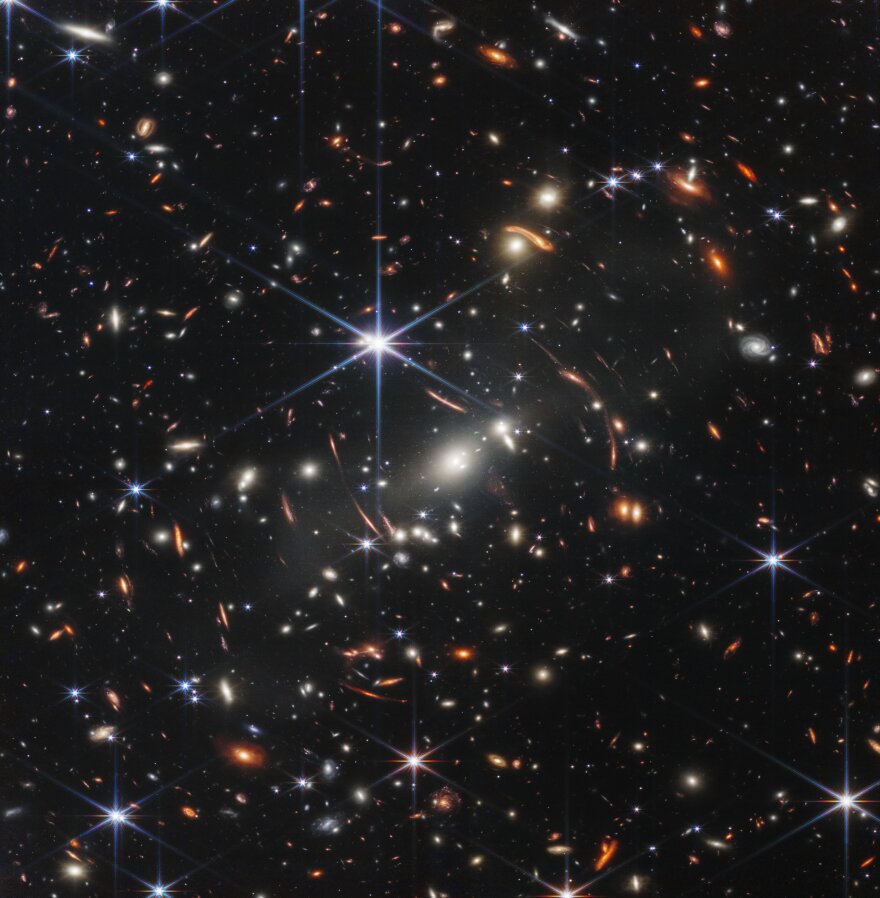 distant galaxies appear as bright glowing spots in this Webb telescope image, with some smeared by gravitational lensing; foreground stars appear bright with six-pointed diffraction spikes, owing to the shape of Webb's mirrors.