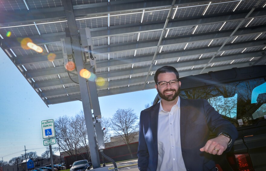 A bearded man wearing a sport coat leans on the edge of a vehicle. He is standing beneath a parking cover that is comprised of solar panels.