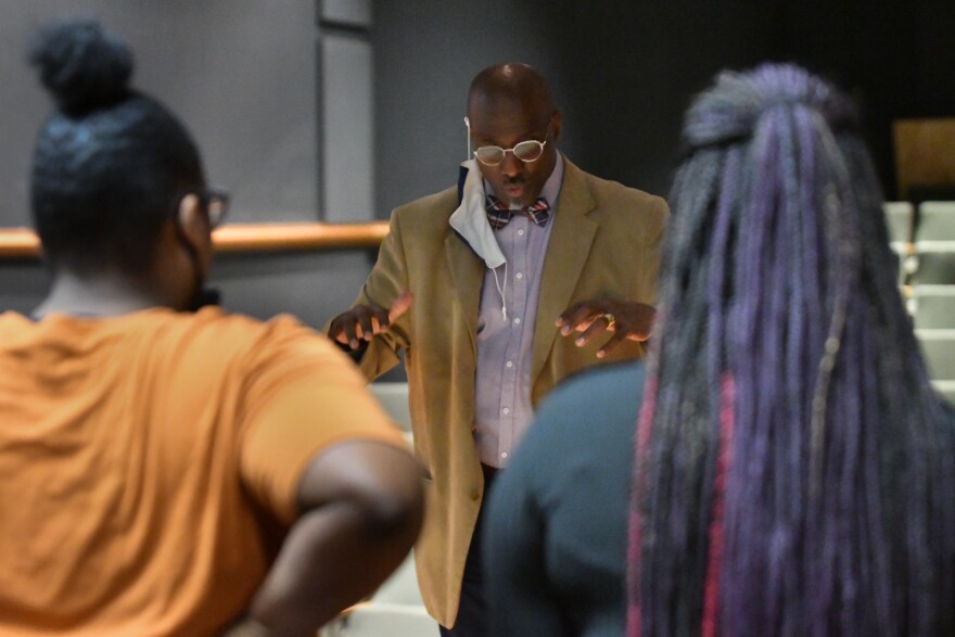 A man, standing in the middle of the frame, gestures with his hands as he conducts high school students during choir practice. In foreground are the backs of two girls who are in the choir.