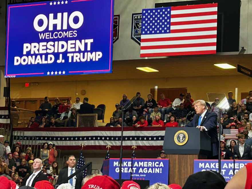 President Donald Trump speaks at a podium in a high school stadium full of supporters, with an american flag behind him
