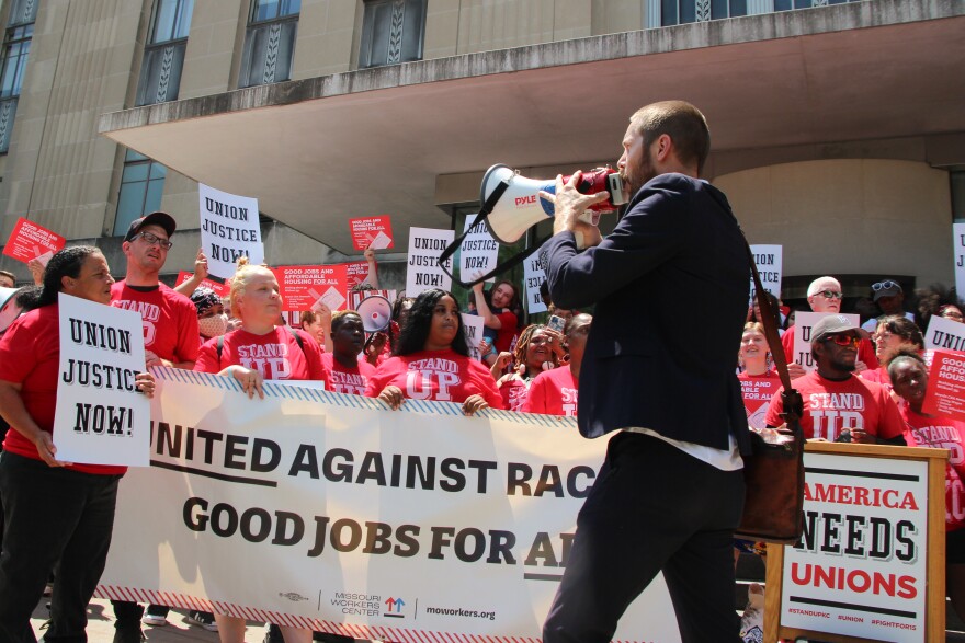  A man in a blue suit holds a megaphone and leads a chant. Behind him is a crowd of people holding signs that advocate for union jobs. They are wearing red shirts that say Stand Up KC and are chanting.