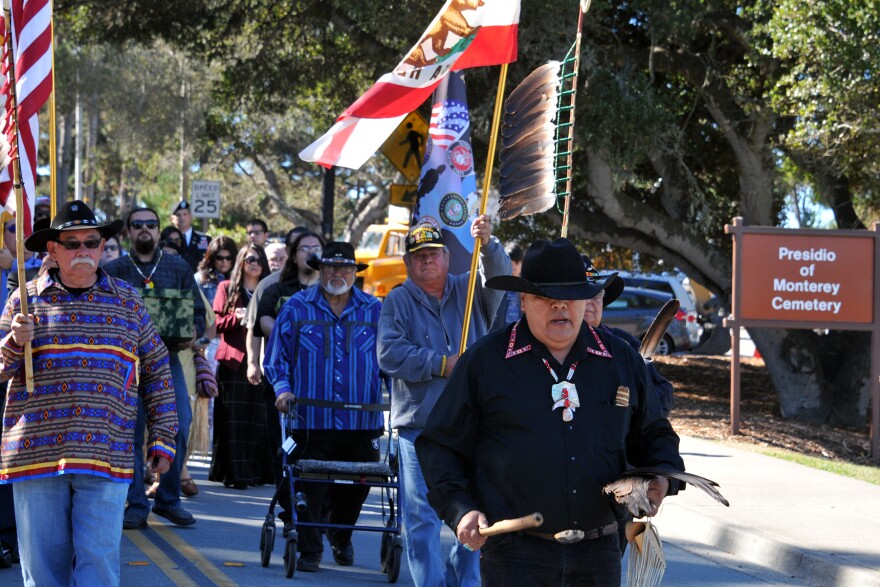 A group of Native Americans and military personnel walks down a street during a funeral procession.