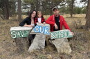 Three people lean against three pieces of wood, with the words "Save Our School."