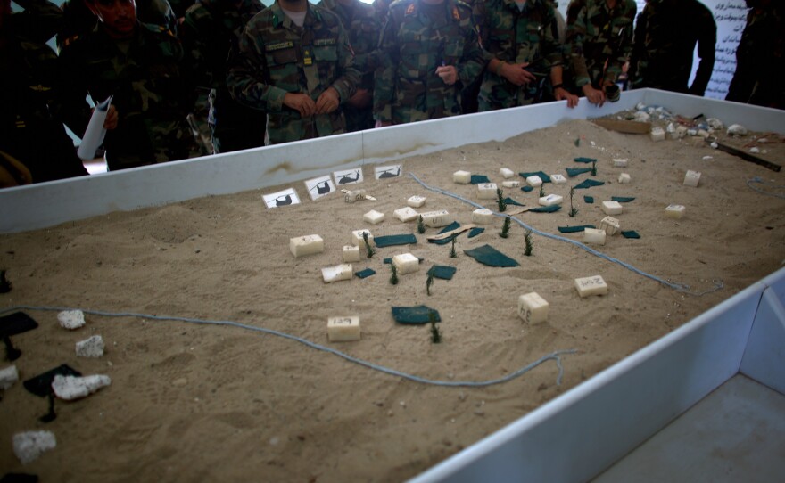 Afghan commandos stand around a sandbox table, studying a model of the village they are about to sweep from Taliban fighters. Styrofoam buildings and green toothpick trees are carefully placed to create a realistic view of the village.
