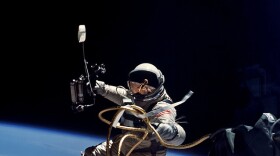 Ed White performs the first U.S. spacewalk. White floats in space with astronaut suit and attached to the shuttle by a cord. Earth is in the background.