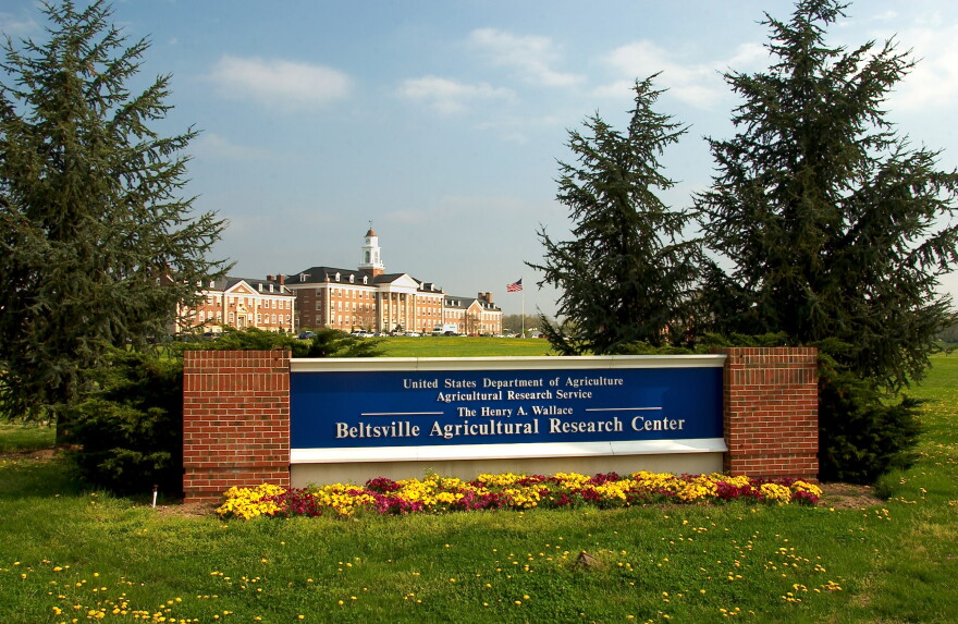 A blue sign reading "Beltsville Agricultural Research Center" sits at the entrance of a research campus, with a large building in the background
