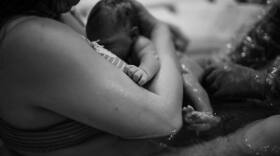 A black and white photo of a mother in a bath tub experiencing a water birth. She holds a newborn baby in her arms. Two hands, a midwife's reach out to the baby.