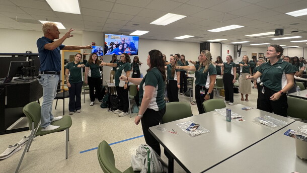 Kevin Spencer stands on a chair with his left arm outstretched to explain a magic trick to a classroom of students at the University of Alabama at Birmingham. The students stand mimicking Spencers position.