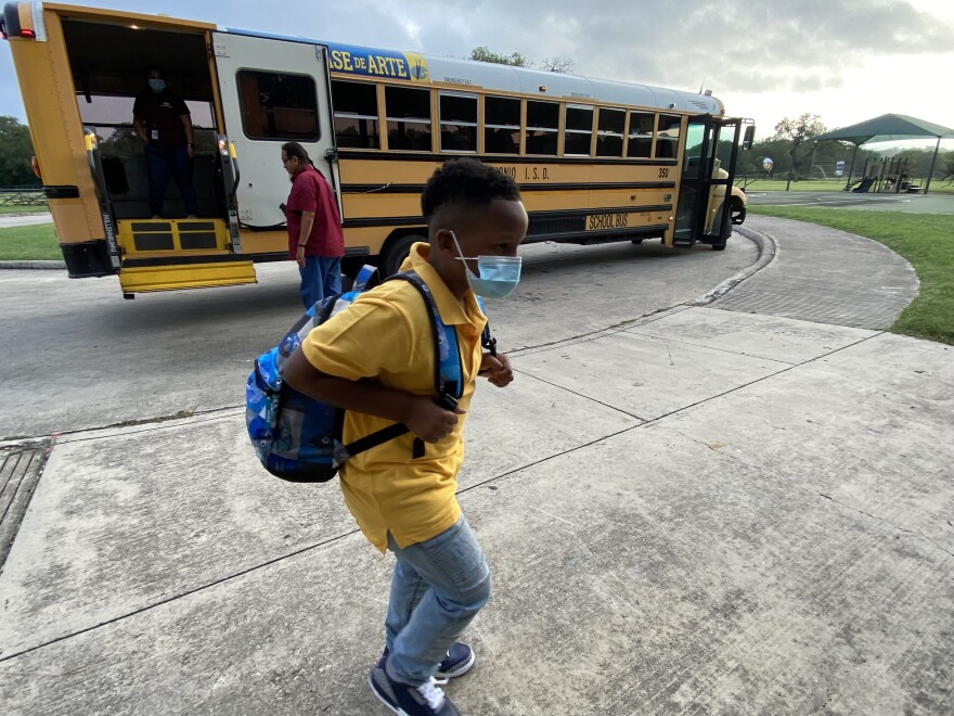 A young San Antonio ISD student walks towards school as a staff member helps a student in a wheelchair off the school bus behind him.