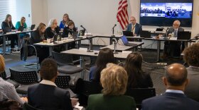A group of people in business attire sit at tables arranged in a U-shape, attending a meeting with lawmakers in a conference room. An American flag is visible, and a video conference is displayed on a screen in the background.