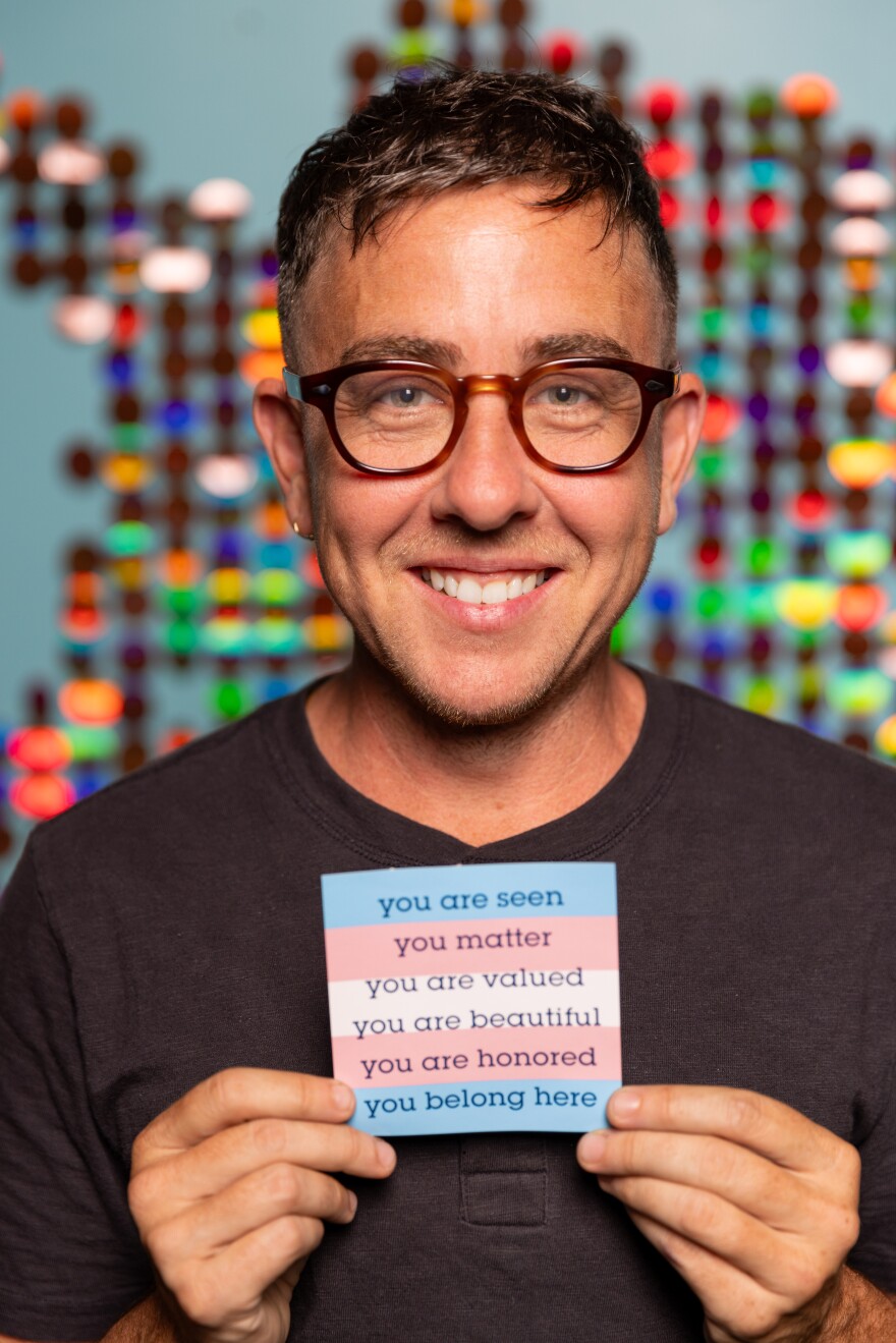 A photo of a smiling person with short brown hair and glasses. The person is holding up a light blue, light pink and white striped card reading "you are seen, you matter, you are valued, you are beautiful, you are honored, you belong here." Blurred in the background is multicolored art.