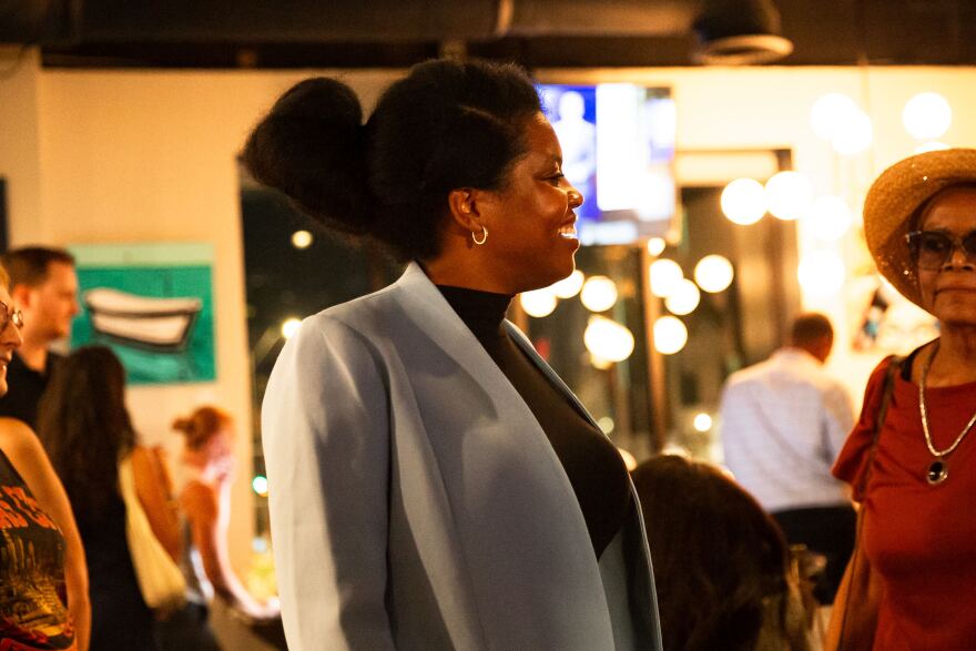 A Black woman in a black turtleneck and gray or light blue blazer stands in profile in a photo at an indoor election watch party. 