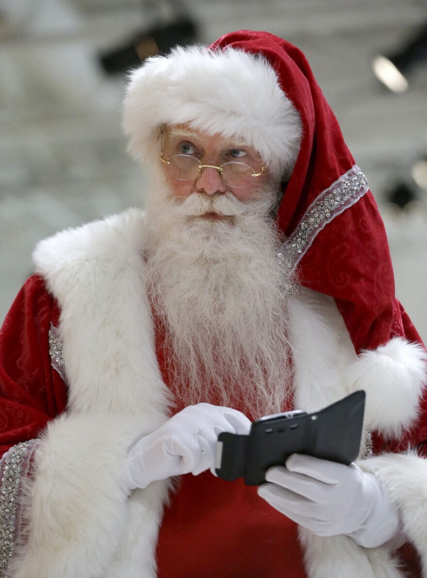 A person dressed as Santa Claus, with a red suit and white fur trim, white gloves, and glasses, is looking up while using a smartphone or tablet device.