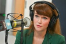 A woman sits inside a radio studio. She is sitting down and talking at a microphone.