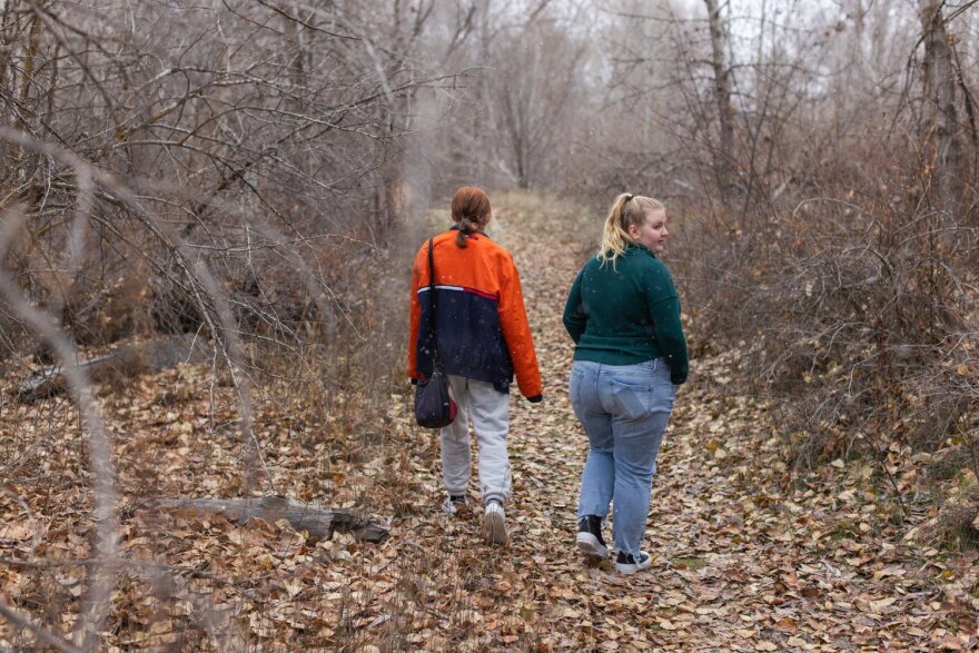 Two teens, one with long blonde hair wearing blue jeans and a green sweater and the other with long red hair and blonde highlights in gray sweats and an orange and black jacket, walking away on a leaf-covered forest path.