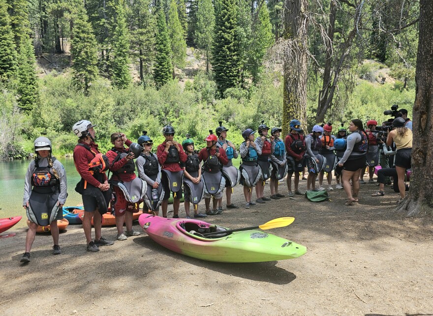 Group of young kayakers in helmets and gear stand in a line beside the Wood River, preparing for their expedition.
