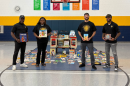 Four adults standing in a school gym holding children's books, next to a mobile book cart and dozens of books spread on the floor.