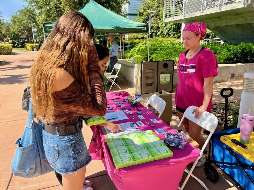 Young women approach a table covered with boxes of emergency contraception. Another woman stands behind the table talking to them.