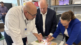 Three people lean over a table, looking closely at a sheet of paper with colored lines.