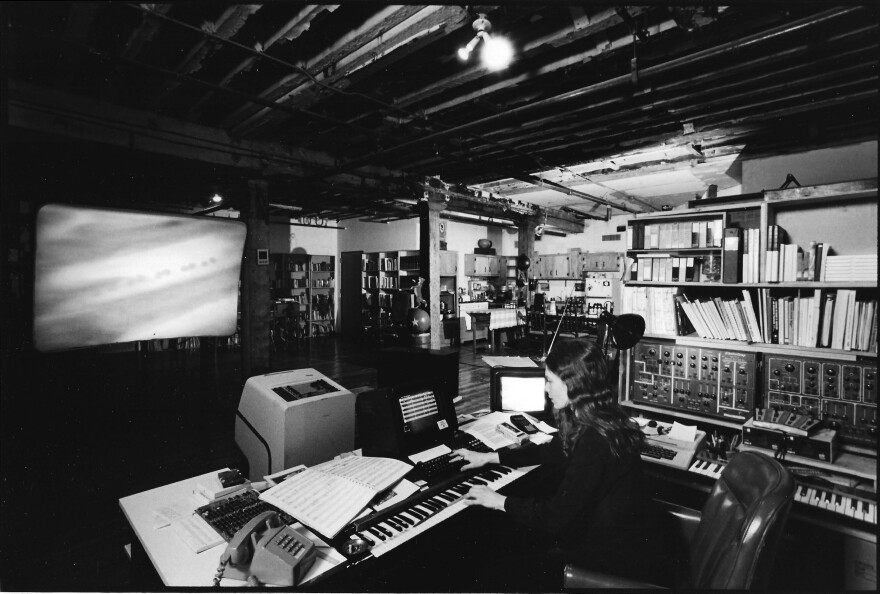 Laurie Spiegel in 1985, with footage of NASA's first Jupiter fly-by projected on the screen in front of her. After twice losing access to tools she used to make her music, Spiegel invented a computer program called Music Mouse that she used to create her 1991 album <em>Unseen Worlds</em>, which was reissued this year.