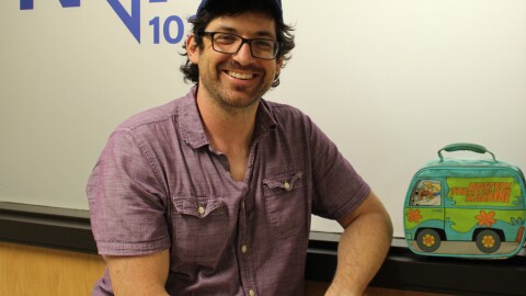 Smiling man wearing a baseball cap seen from the waist up in front of a frosted window that says The North 103.3 FM. There is a lunch box shaped like the Scooby Doo Mystery Machine to his right.