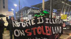 People, many with umbrellas, stand near a construction site facing the street. Some are holding a large banner that says "No One is Illegal on Stolen Land - #stop the sweeps"