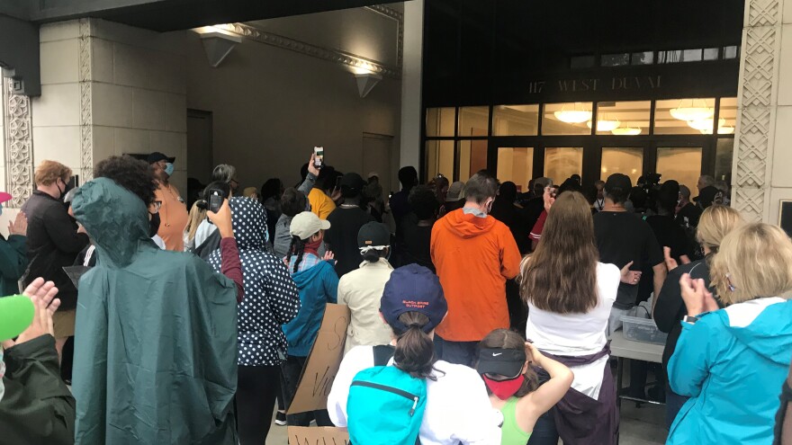 People in a crowd, many in masks, looking toward the entrance of city hall, where elected officials are speaking