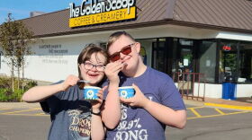 A woman and man stand together posing for a photo in front of The Golden Scoop Coffee and Creamery. Each person has a cup and spoon in their hand. 