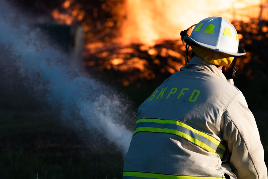 A firefighter wearing a helmet and grey jacket with yellow stripes and the letters BKPFD uses a hose to spray water on a large fire