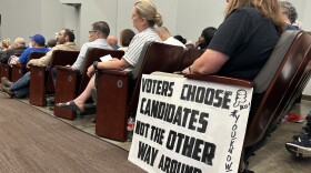 A photo of people listening to a speaker in auditorium seats. A sign is propped up against a chair that says, "Voters choose candidates, not the other way around."