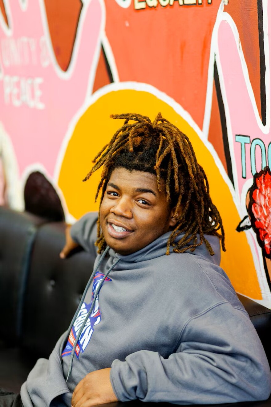A young man with highlighted brown and black dreadlocks sits on a dark leather couch, smiling at the camera. He wears a grey hooded sweatshirt with a blue and red graphic logo on the chest. Behind him is a brightly colored mural featuring shades of orange, yellow, and pink with text fragments like "PEACE" visible on the left side.