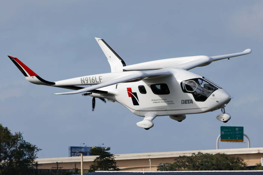 A white s electric aircraft flies above a highway, with its wings, propellers and landing gear visible against a clear blue sky.