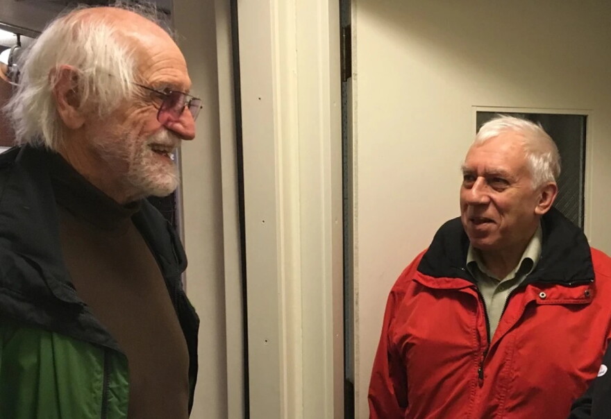 Man in a green coat and brown sweater standing in a doorway speaking to a man in a red coat in front of an interior door.